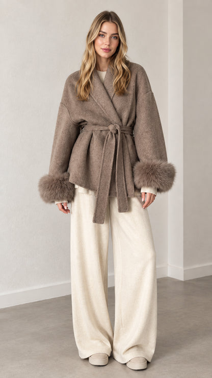 Front view of model wearing a taupe belted wrap coat with faux fur cuffs and white trousers